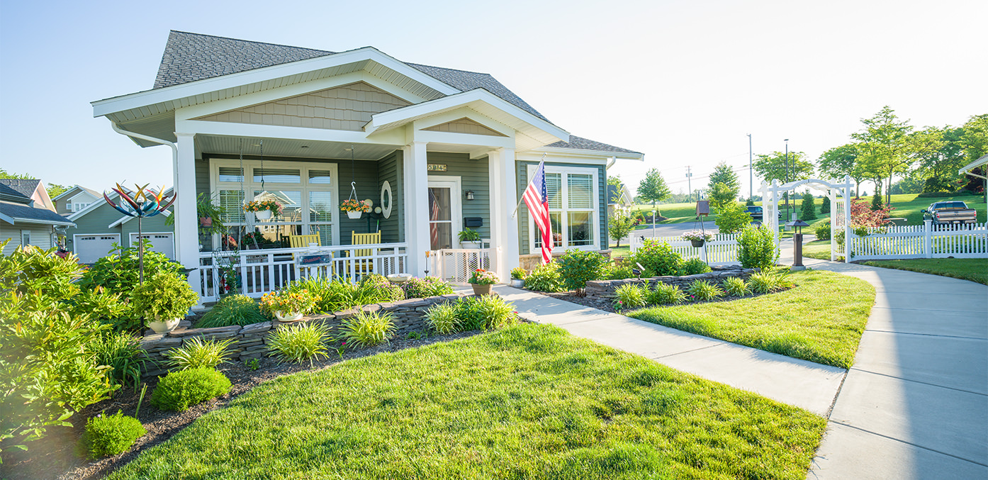 Sun illuminating a green residential house front view that has manicured flower beds and yard, and an American flag hanging from the front of the house